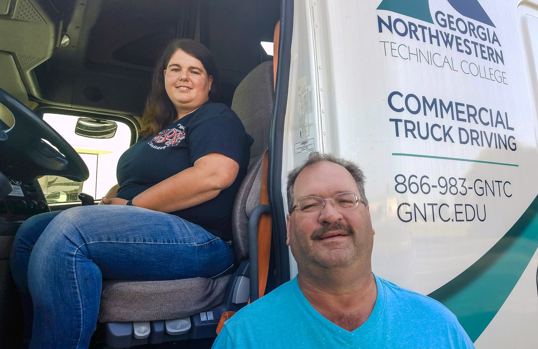 Gordon County, Georgia’s Ashley Petty poses from the cab of a Georgia Northwestern Technical College Commercial Truck Driving program tractor-trailer. Standing in the foreground on a hot day at the college’s truck driving facility is Georgia Northwestern Technical College Program Director Robert Browder.